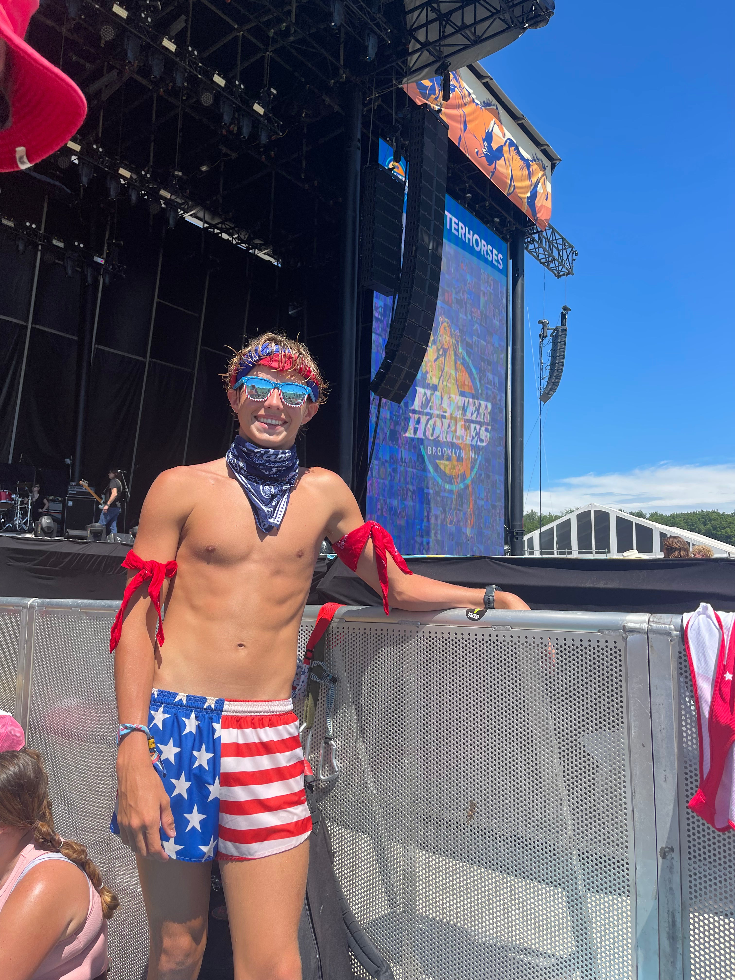 Men's Flag 1 Elite Split Shorts in a bold red, white, and blue American flag print with stars and stripes design, worn by a young man at an outdoor music festival.