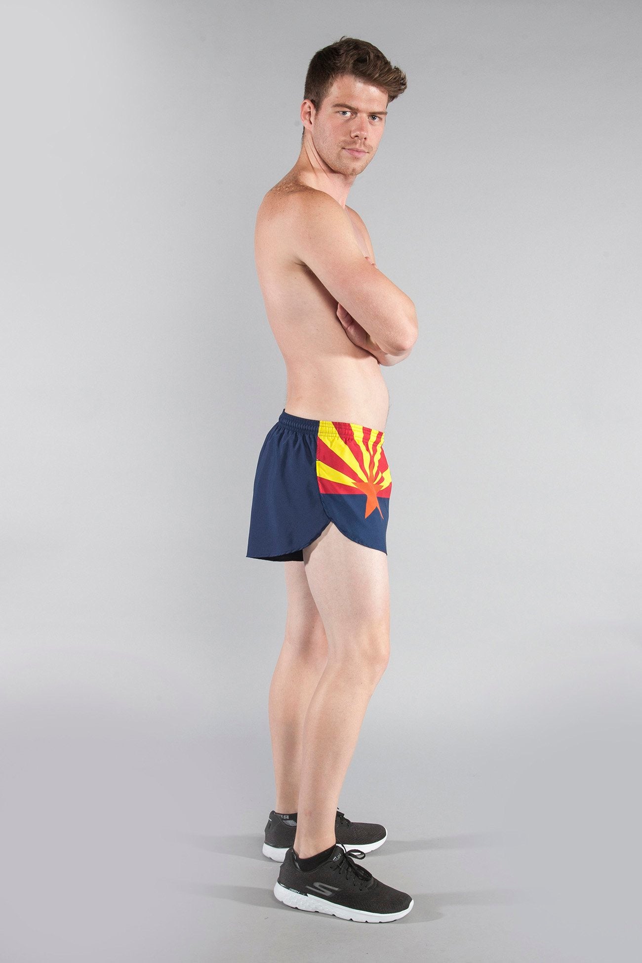 Men's Flag 1" Elite Split Shorts - Arizona, featuring the Arizona state flag design with navy blue, red, yellow, and orange star burst pattern, modeled by a shirtless male wearing black Skechers sneakers, arms crossed, side profile view.