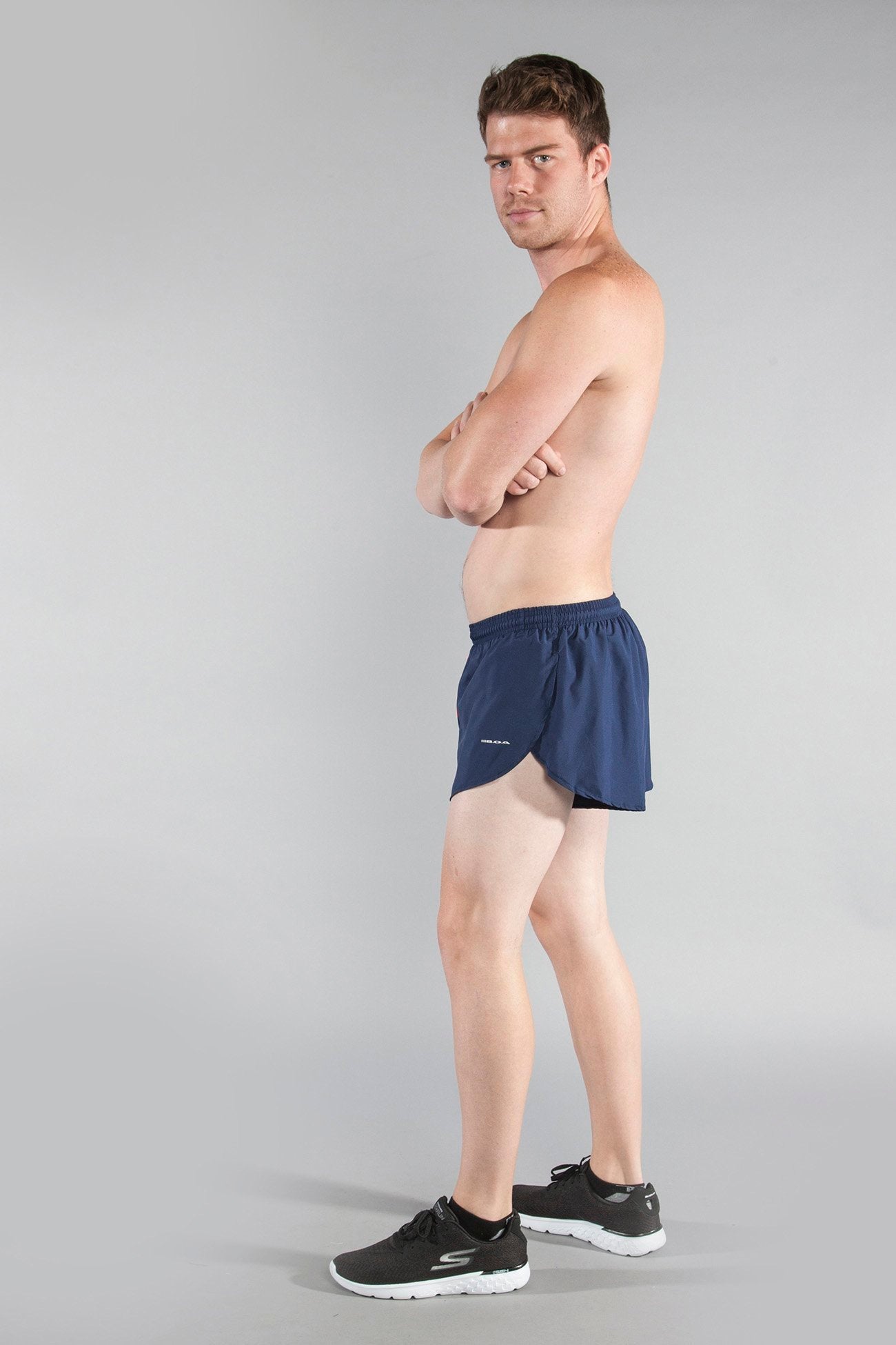Men's Flag 1" Elite Split Shorts Arizona in navy blue, modeled by a shirtless man with arms crossed, wearing black Skechers sneakers, shown from a side angle against a light gray background.