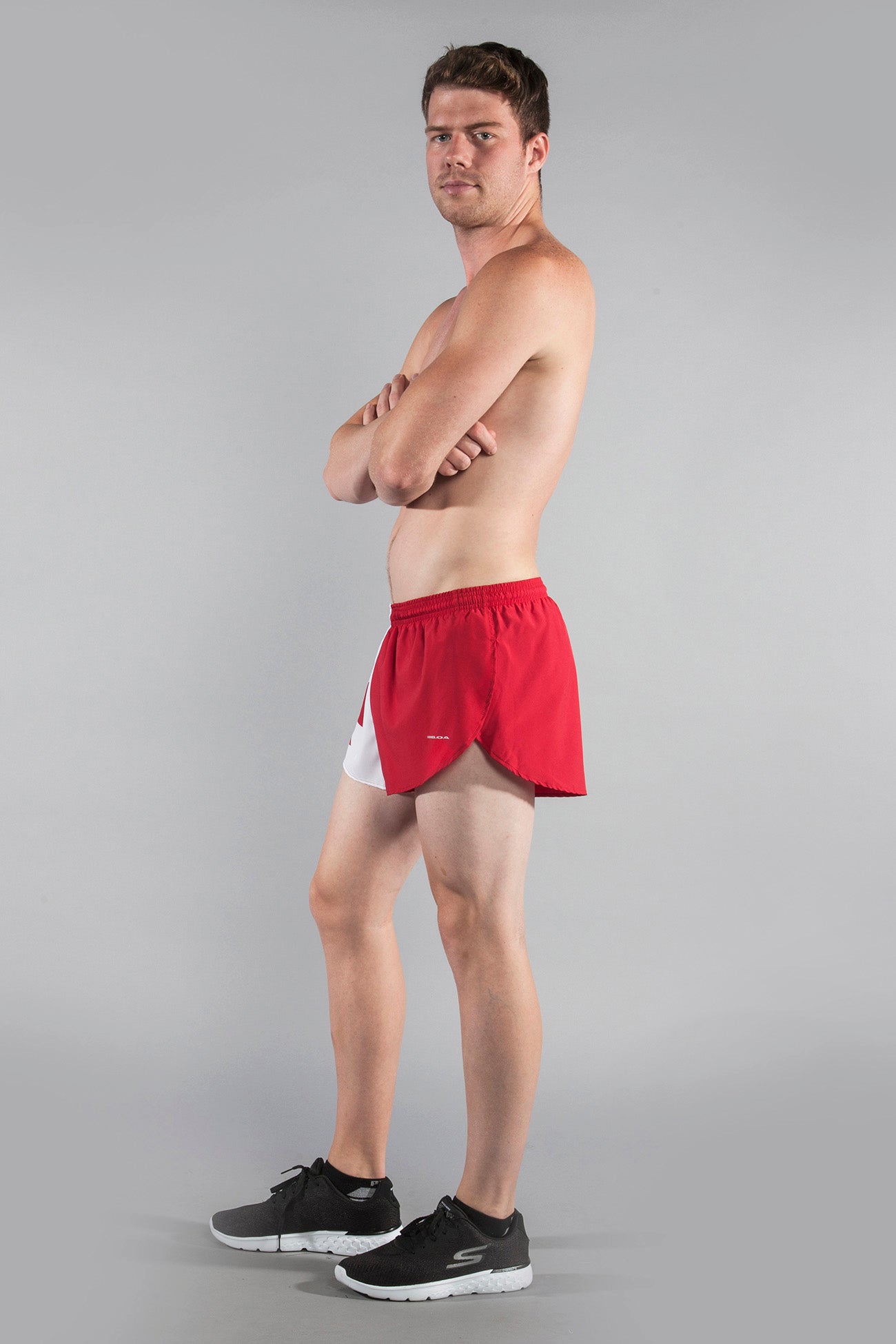 Men's Flag 1" Elite Split Shorts in red and white Canada flag design, modeled by a shirtless man with arms crossed, paired with black Skechers sneakers, shown from a side angle against a gray background.