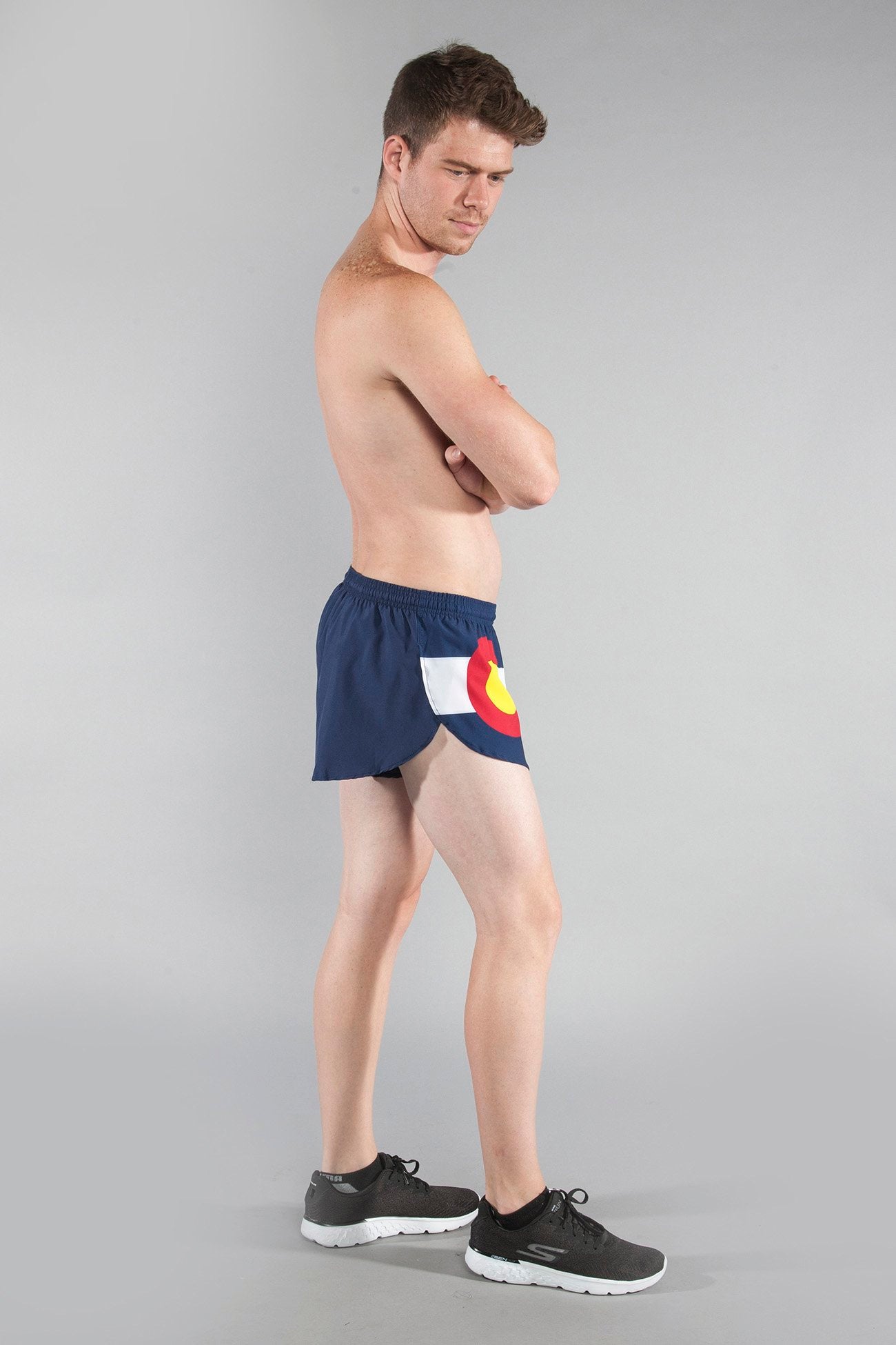 Men's Flag 1" Elite Split Shorts in navy blue featuring the Colorado state flag emblem, modeled by a shirtless man paired with black Skechers sneakers against a gray background.