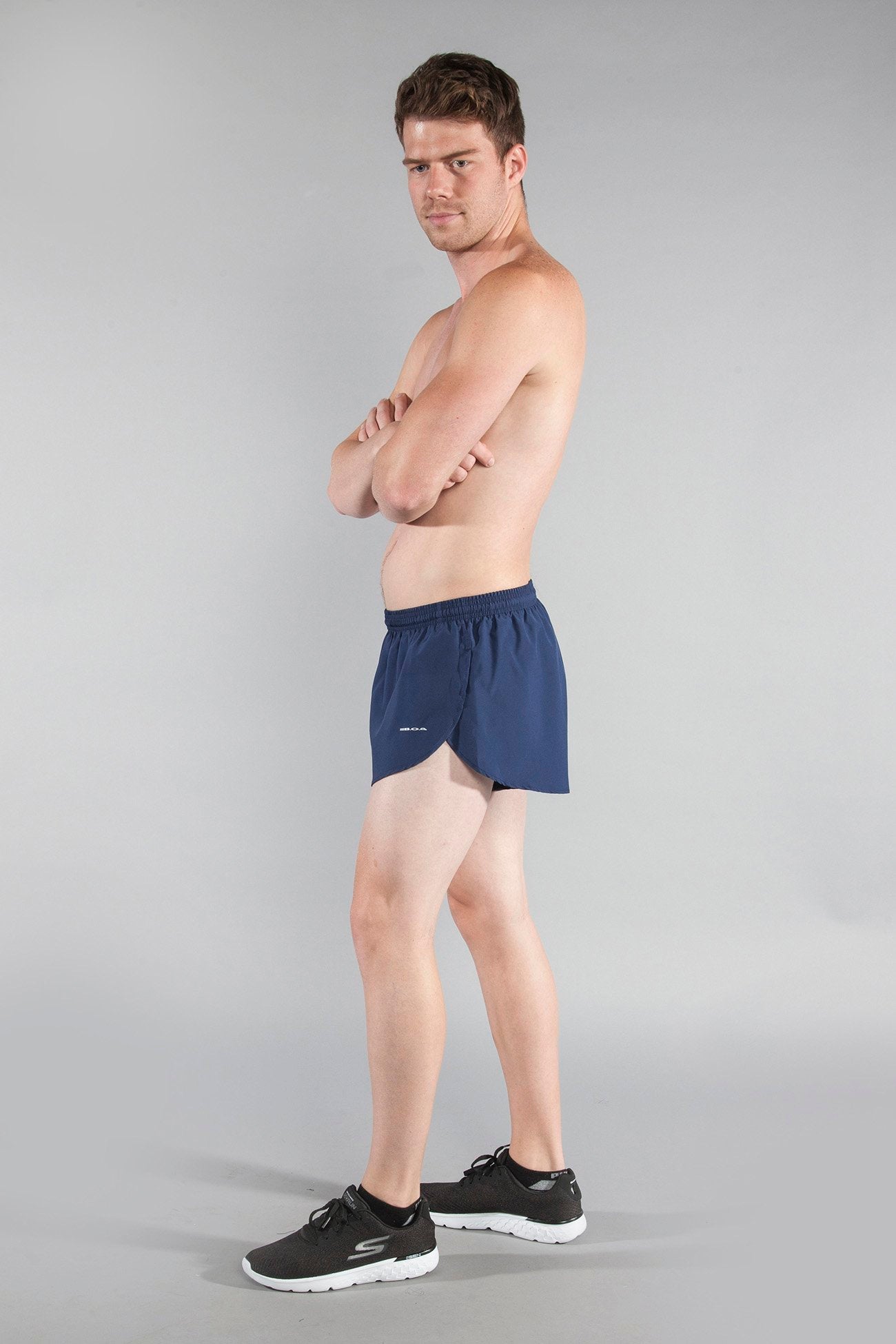 Men's Flag 1" Elite Split Shorts in navy blue, modeled by a shirtless man with arms crossed, paired with black Skechers sneakers, shown from a side angle against a gray background.