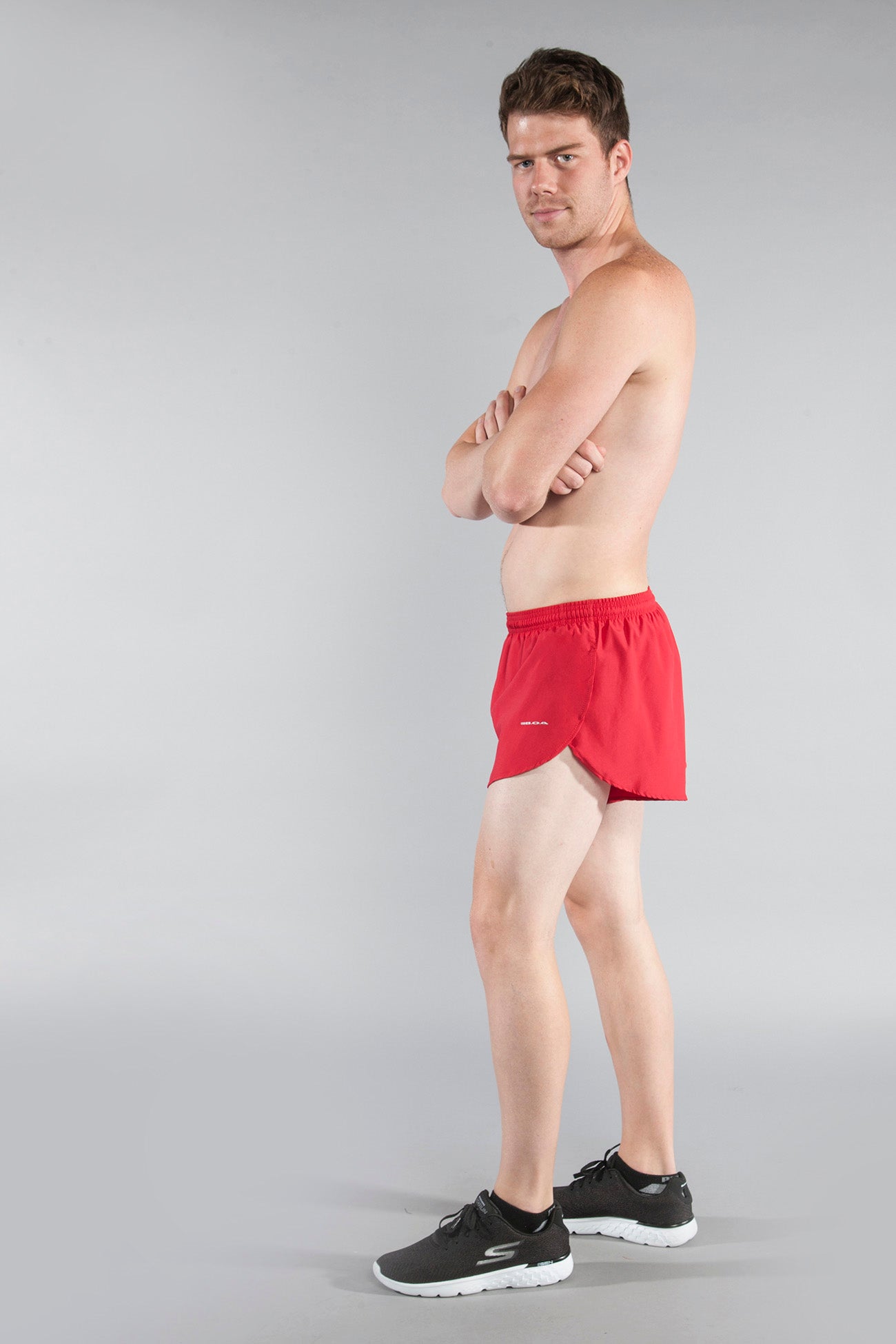 Men's Flag 1" Elite Split Shorts in red worn by a shirtless male model with arms crossed, shown from a side angle, paired with black Skechers sneakers — Florida style athletic running shorts with a split hem design.