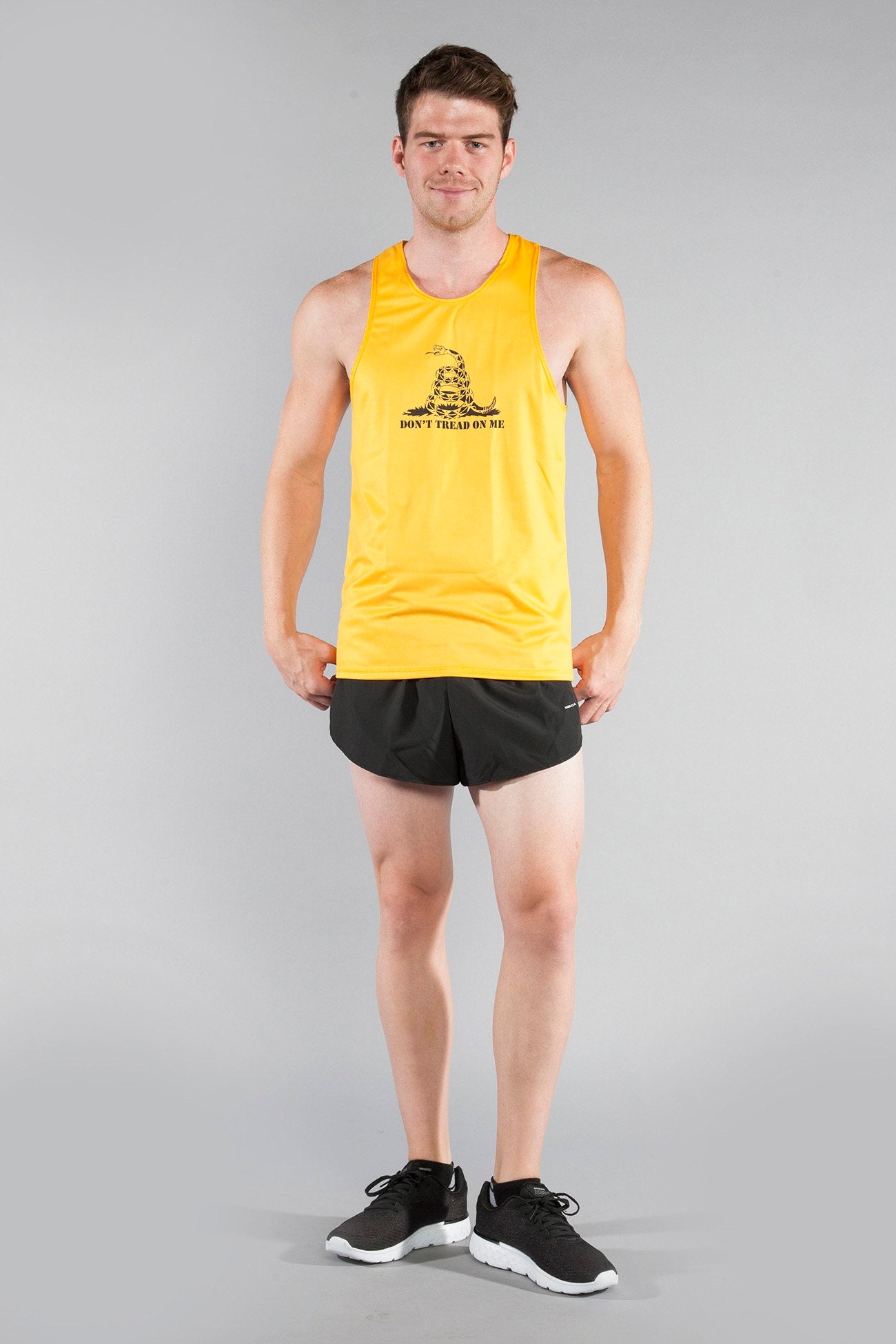 Man wearing Men's Competitor Lite Printed Singlet in yellow featuring a coiled rattlesnake and Don't Tread on Me text, paired with black running shorts and black athletic shoes, standing against a gray background.