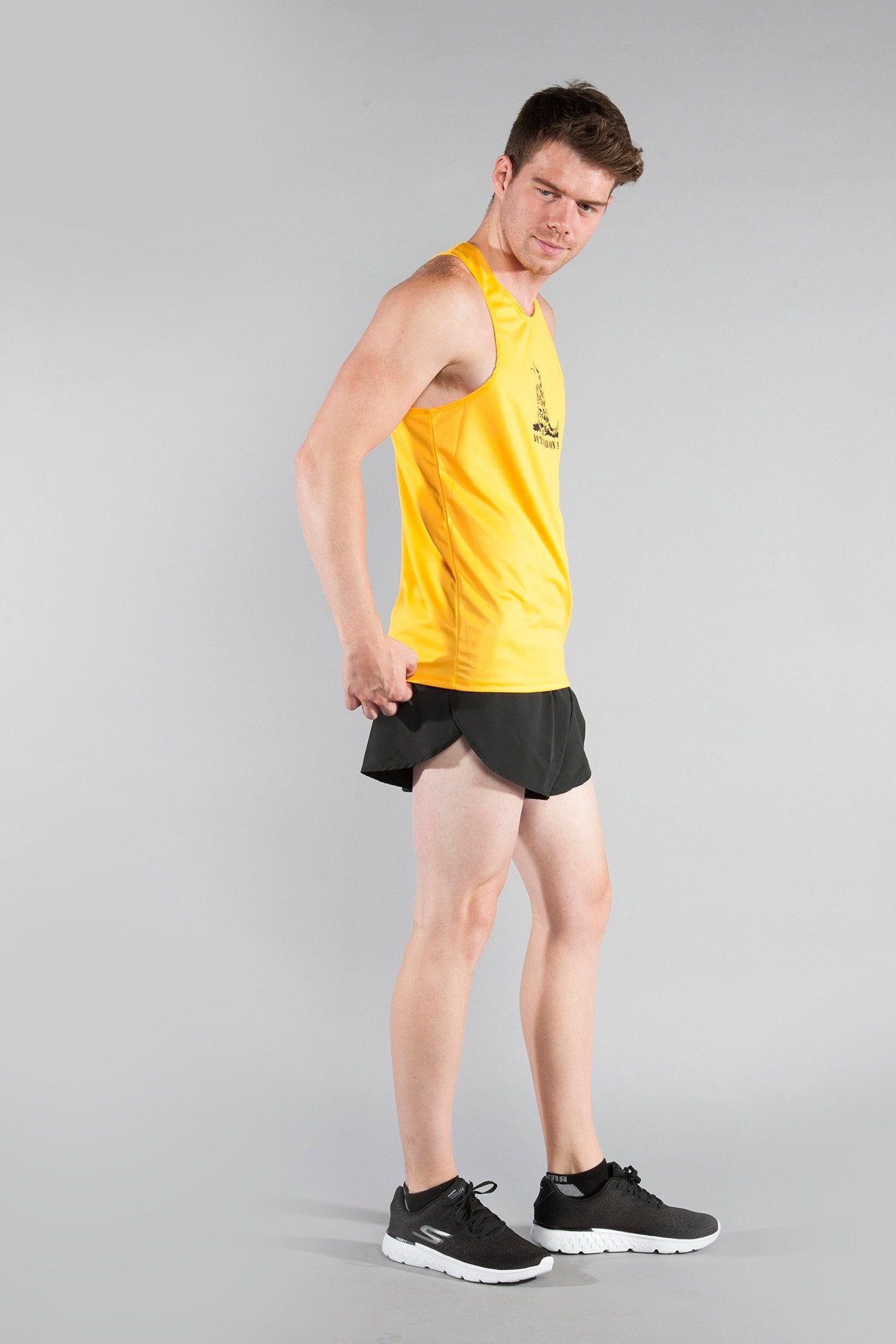 Man modeling the Men's Competitor Lite Printed Singlet in golden yellow featuring a Don't Tread on Me snake graphic on the chest, paired with black running shorts and black Skechers sneakers, shown in a three-quarter side pose against a gray background.